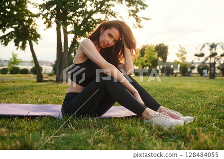 Sitting on the yoga mat against sunlight. Beautiful young woman in sportive clothes is in the park at sunny daytime 128484055