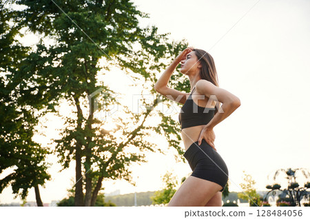 Exercises, against tree and sky. Beautiful young woman in sportive clothes is in the park at sunny daytime 128484056