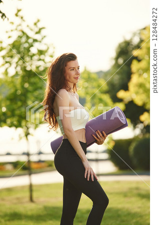 With yoga mat. Beautiful young woman in sportive clothes is in the park at sunny daytime 128484272