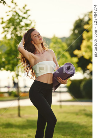 Holding yoga mat in hands. Beautiful young woman in sportive clothes is in the park at sunny daytime 128484274