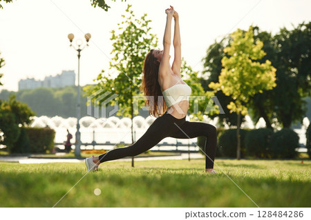 Warrior yoga asana. Beautiful young woman in sportive clothes is in the park at sunny daytime Warrior yoga asana. Beautiful young woman in sportive clothes is in the park at sunny daytime 128484286