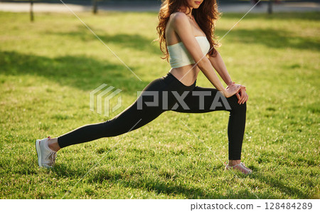 On green grass, stretching the legs. Beautiful young woman in sportive clothes is in the park at sunny daytime 128484289