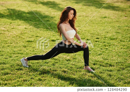 On green grass, stretching the legs. Beautiful young woman in sportive clothes is in the park at sunny daytime 128484290