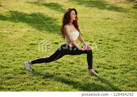 On green grass, stretching the legs. Beautiful young woman in sportive clothes is in the park at sunny daytime 128484292