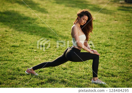 On green grass, stretching the legs. Beautiful young woman in sportive clothes is in the park at sunny daytime 128484295