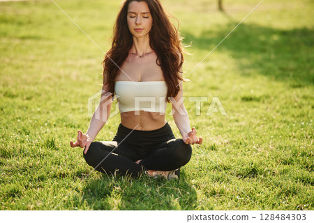 Meditating in lotus pose. Beautiful young woman in sportive clothes is in the park at sunny daytime Meditating in lotus pose. Beautiful young woman in sportive clothes is in the park at sunny daytime 128484303
