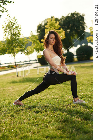 Doing fitness. Beautiful young woman in sportive clothes is in the park at sunny daytime 128484321