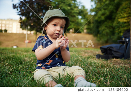 Cute little boy in hat that is with ears is sitting on the grass in the park at daytime 128484400