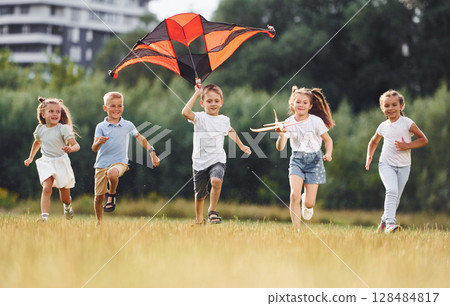 Running together. Group of kids are running and playing with kite on green field 128484817