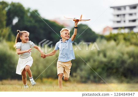 Holding each other by hands. Boy and girl are playing with toy plane on the green field 128484947