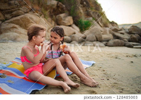 Joyful sisters enjoying snacks on the sandy beach at sunset 128485003