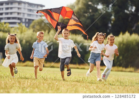 Running together. Group of kids are running and playing with kite on green field 128485172