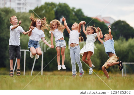 Holding each other by hands and jumping. Kids are having fun on the field at daytime together 128485202
