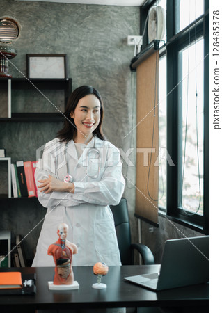 Female doctor in a modern office setting, wearing a white coat, with anatomical models and a laptop on the desk. 128485378
