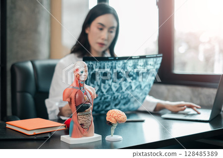 A medical professional reviews X-ray images in a contemporary office setting, featuring anatomical models of the human body and brain. 128485389