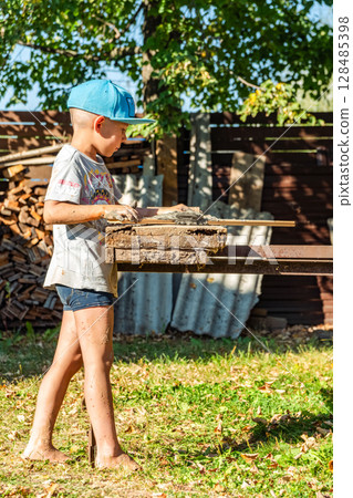 Young boy in a cap cheerfully creates with clay, standing barefoot and covered in dirt, near a rustic wooden yard table 128485398