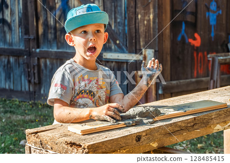 Little boy wearing a cap is focused on shaping clay in the yard of a rustic village house, enjoying a sunny day outdoors 128485415