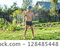 Joyful boy in a cap enjoys splashing water from a hose, playfully cleaning himself in a vibrant garden under bright sunshine 128485448