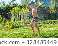 Joyful boy in a cap enjoys splashing water from a hose, playfully cleaning himself in a vibrant garden under bright sunshine 128485449