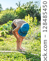 Joyful boy in a cap enjoys splashing water from a hose, playfully cleaning himself in a vibrant garden under bright sunshine 128485450