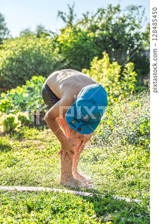 Joyful boy in a cap enjoys splashing water from a hose, playfully cleaning himself in a vibrant garden under bright sunshine 128485450
