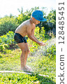 Joyful boy in a cap enjoys splashing water from a hose, playfully cleaning himself in a vibrant garden under bright sunshine 128485451