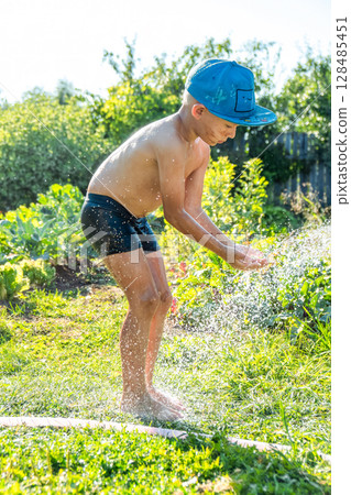 Joyful boy in a cap enjoys splashing water from a hose, playfully cleaning himself in a vibrant garden under bright sunshine Joyful boy in a cap enjoys splashing water from a hose, playfully cleaning himself in a vibrant garden under bright sunshine 128485451