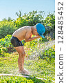 Joyful boy in a cap enjoys splashing water from a hose, playfully cleaning himself in a vibrant garden under bright sunshine 128485452