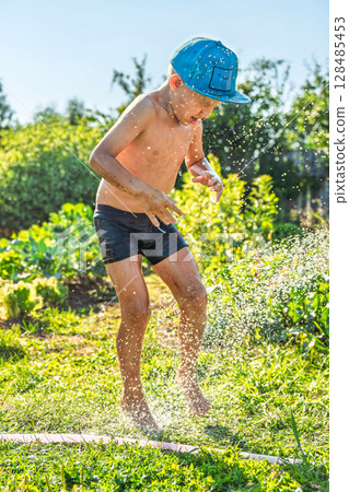 Joyful boy in a cap enjoys splashing water from a hose, playfully cleaning himself in a vibrant garden under bright sunshine 128485453