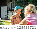 Little boy, dirty and smiling, faces his grandmother who appears to be scolding him in a country yard, on a sunny day 128485721