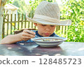 Young boy wearing a hat savours a bowl of soup, sitting comfortably at a rustic wooden table in a sunny garden 128485723