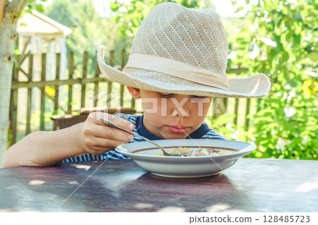 Young boy wearing a hat savours a bowl of soup, sitting comfortably at a rustic wooden table in a sunny garden Young boy wearing a hat savours a bowl of soup, sitting comfortably at a rustic wooden table in a sunny garden 128485723