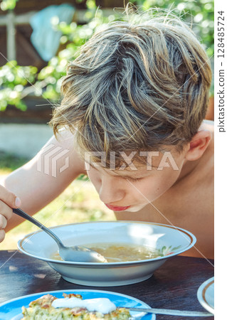 Boy with thick hair enjoys lunch at a wooden table in a sunny country garden setting 128485724
