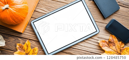A digital tablet with copy space surrounded by autumn leaves, a pumpkin, and a phone on a wooden table 128485750