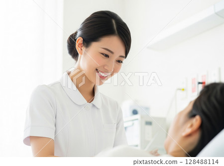 A female nurse in a white coat gently speaks to a patient at the bedside 128488114