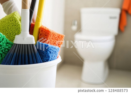 Person is standing in the WC holding a blue bucket full of chemicals, cleaning supplies and facilities for cleansing. Clean toilet. Neat restroom. House cleanliness, service. White ceramic toilet bowl 128488573