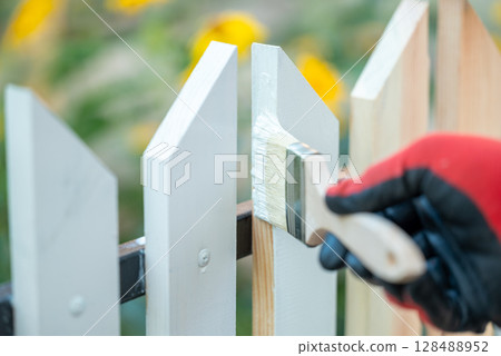 Close up of man hand in protective glove painting fence in white color. Painting new wooden fence with a brush in white color Close up of man hand in protective glove painting fence in white color. Painting new wooden fence with a brush in white color 128488952