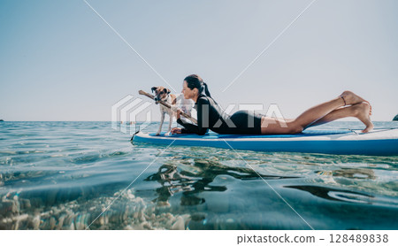 Woman Dog Paddleboarding Ocean - A woman and her dog paddleboard on a clear blue ocean on a sunny day. Woman Dog Paddleboarding Ocean - A woman and her dog paddleboard on a clear blue ocean on a sunny day. 128489838