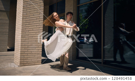 Joyful woman dancing in arms of partner on urban street with sunlit reflection in glass wall 128491458