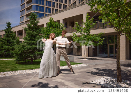 Smiling young couple dancing outdoors in sunlight near urban greenery and modern residential building 128491468