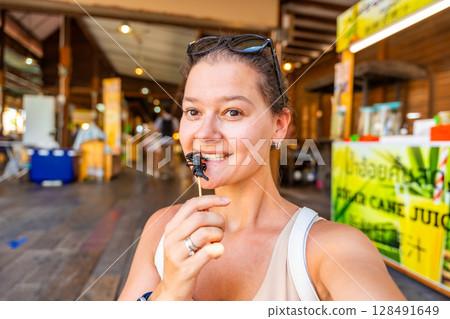 Young woman tourist tasting fried scorpion on a skewer bought in floating open air market on the pond in Pattaya, Thailand 128491649