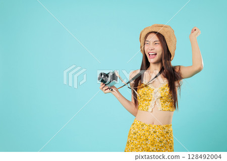 Happy Asian tourist woman holding camera ready for summer travel, she young excited and positive, raising fists in cheerful celebration of her travel holiday, headshots isolated studio blue background 128492004