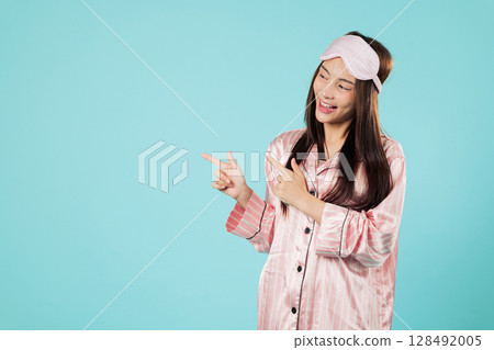 Happy young asian woman pointing wearing pajama, sleep eye mask pointing her finger to an empty space perfect for a nap at home, copy space area for ads, headshots isolated studio blue background 128492005