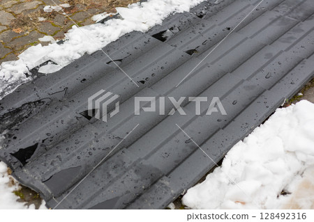 Black roofing panels are placed on the ground amidst patches of snow, indicating recent installation and winter weather conditions Black roofing panels are placed on the ground amidst patches of snow, indicating recent installation and winter weather conditions 128492316