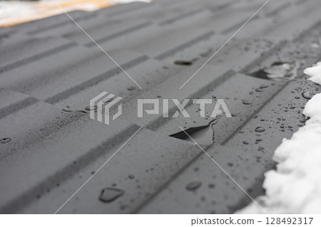 Melting snow creates small pools of water on the surface of newly installed metal roofing, showcasing the material and winter elements Melting snow creates small pools of water on the surface of newly installed metal roofing, showcasing the material and winter elements 128492317