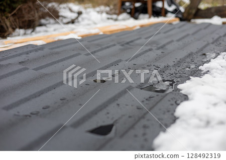 Metal roofing covered partly by fresh snow while water droplets gather on its surface in a residential backyard setting Metal roofing covered partly by fresh snow while water droplets gather on its surface in a residential backyard setting 128492319