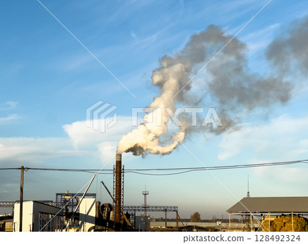 A large smokestack emits white smoke into the blue sky, displaying industrial activity in a manufacturing area during the afternoon A large smokestack emits white smoke into the blue sky, displaying industrial activity in a manufacturing area during the afternoon 128492324