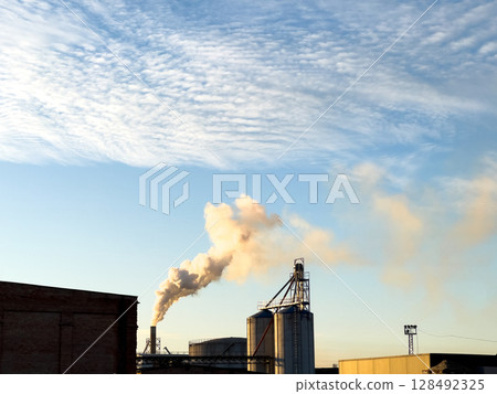 Industrial facility releasing smoke into the air at sunset, showcasing the interplay between urban development and nature Industrial facility releasing smoke into the air at sunset, showcasing the interplay between urban development and nature 128492325