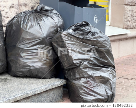 Black garbage bags are piled on the sidewalk beside a public trash can in a busy urban setting, capturing daily waste disposal 128492332