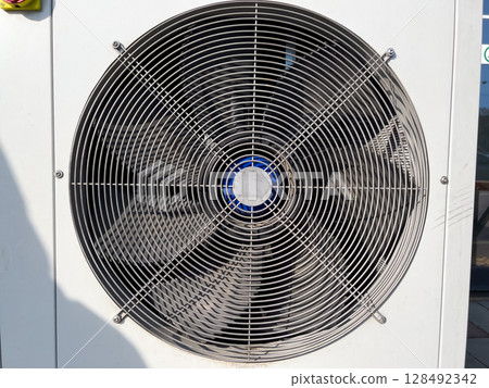 A large ventilation fan with metal grill spins rapidly against a sunny backdrop, highlighting its role in air circulation for a warehouse A large ventilation fan with metal grill spins rapidly against a sunny backdrop, highlighting its role in air circulation for a warehouse 128492342